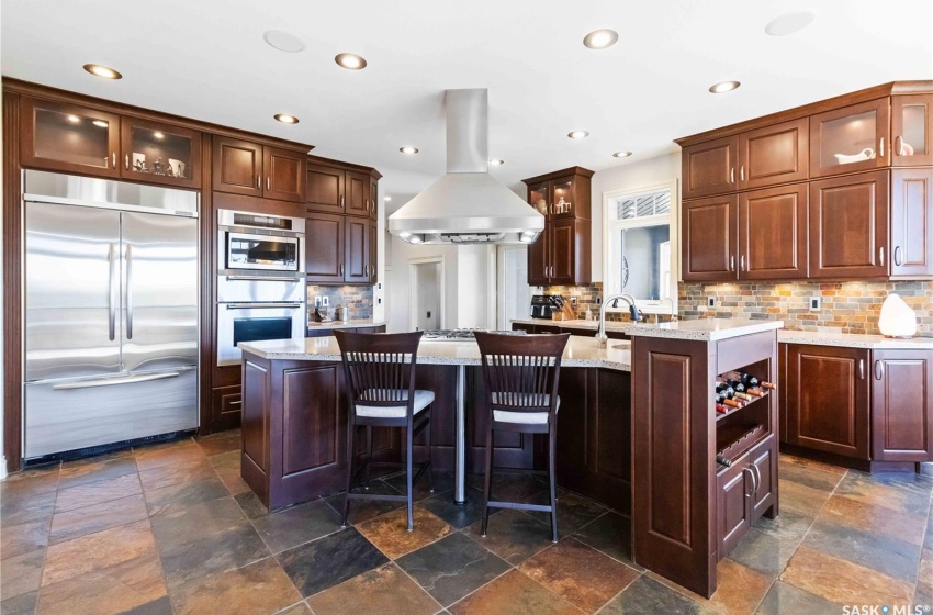 Kitchen with island exhaust hood, appliances with stainless steel finishes, backsplash, light stone countertops, and a center island with sink