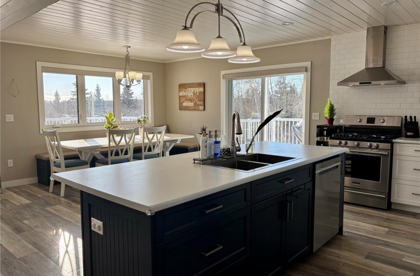 Kitchen with wall chimney range hood, sink, a kitchen island with sink, hanging light fixtures, and stainless steel appliances